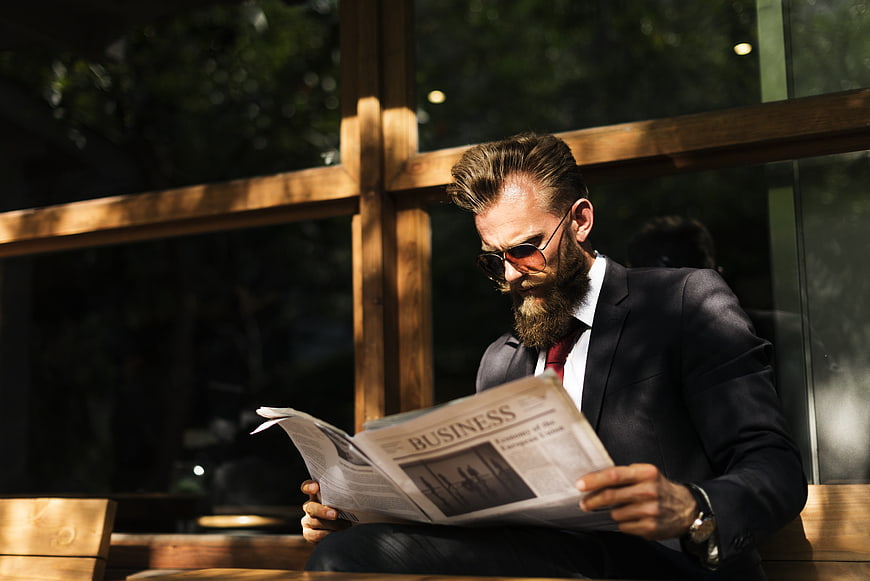 Bearded businessman in suit reading business newspaper in sunlit cafe