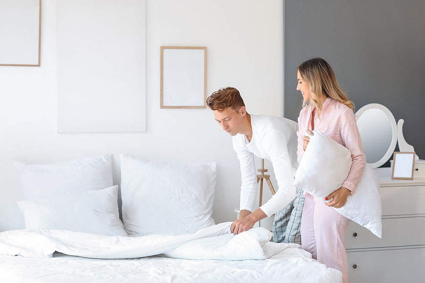 Happy young couple making bed in bright modern bedroom