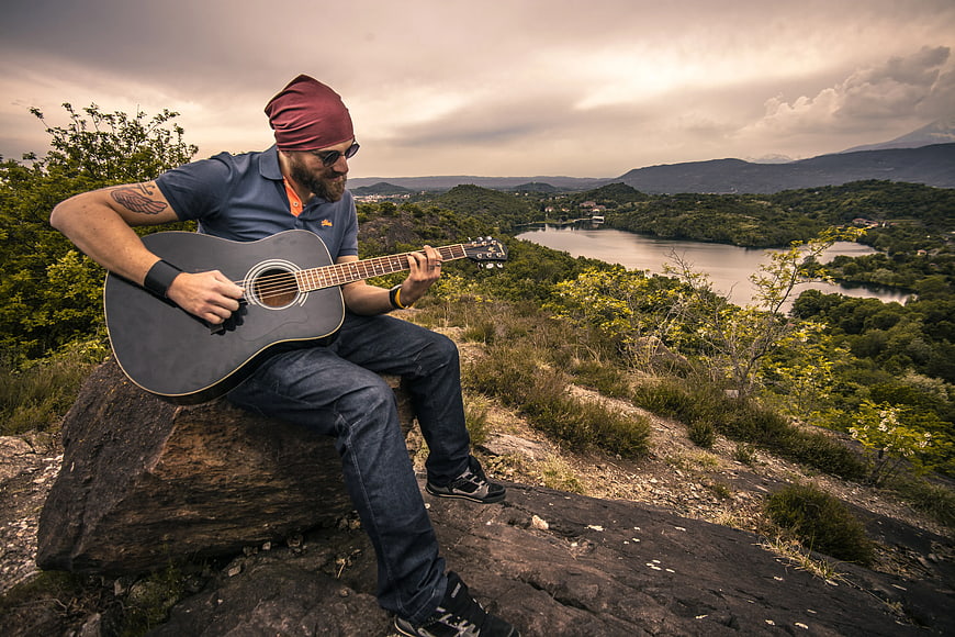 Man playing acoustic guitar on rocky hilltop overlooking lake