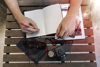 Overhead view of hands writing in notebook with camera and tablet on wooden table