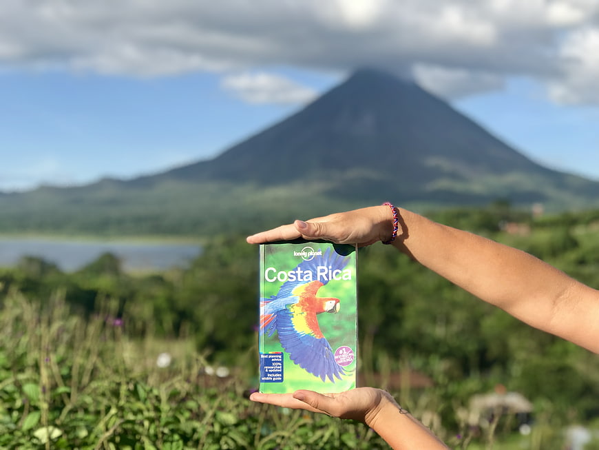Hands holding Lonely Planet Costa Rica guidebook with volcano background
