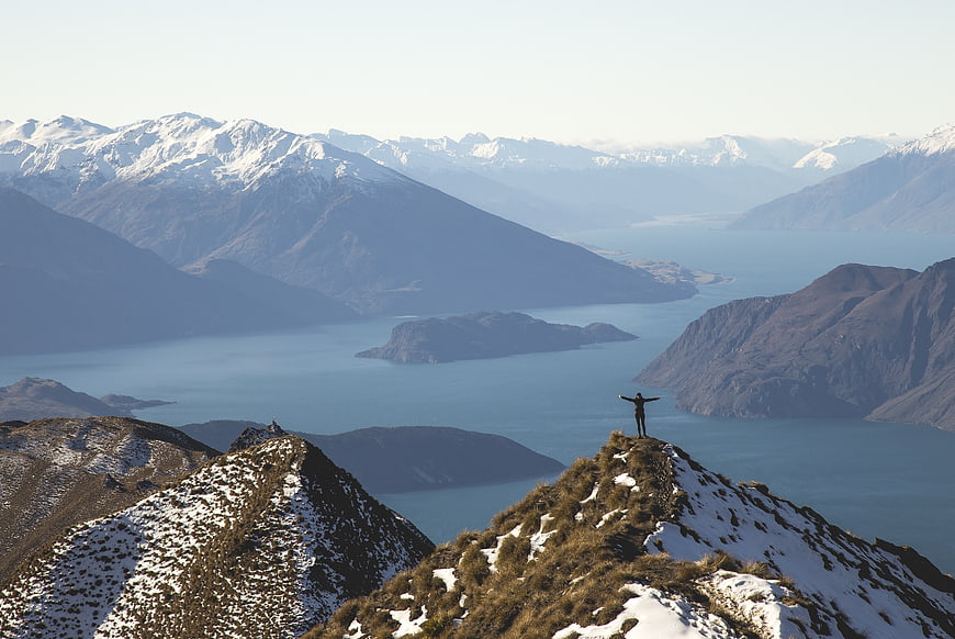 Hiker standing on snowy mountain ridge overlooking lake and peaks