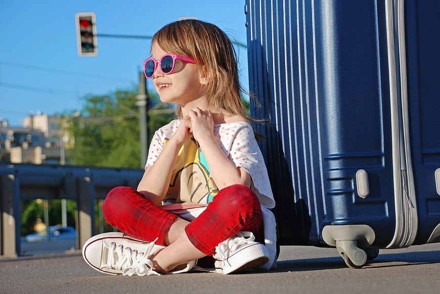 Cute young girl with pink sunglasses sitting next to blue suitcase