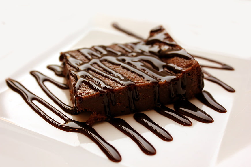 Square chocolate brownie with chocolate drizzle on white plate