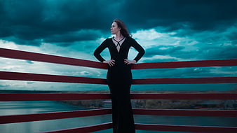 Woman in black dress leaning on red bridge railing against teal sky