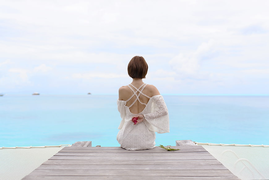 Woman in white dress sitting on pier facing turquoise ocean