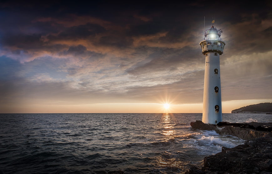 White Lighthouse On Rocky Coast At Sunset With Dramatic Clouds