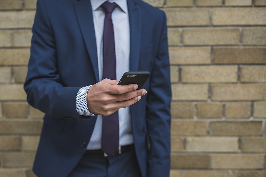 Businessman in Blue Suit Using Smartphone Against Brick Wall