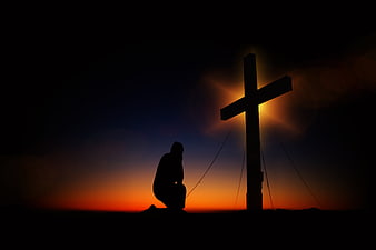 Silhouette of person kneeling in prayer before cross at sunset