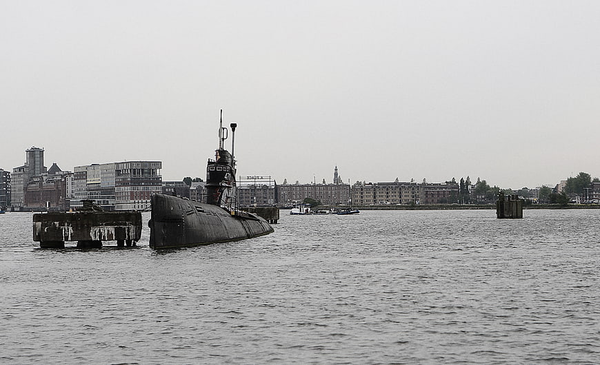 Black submarine docked in Amsterdam harbor with city skyline