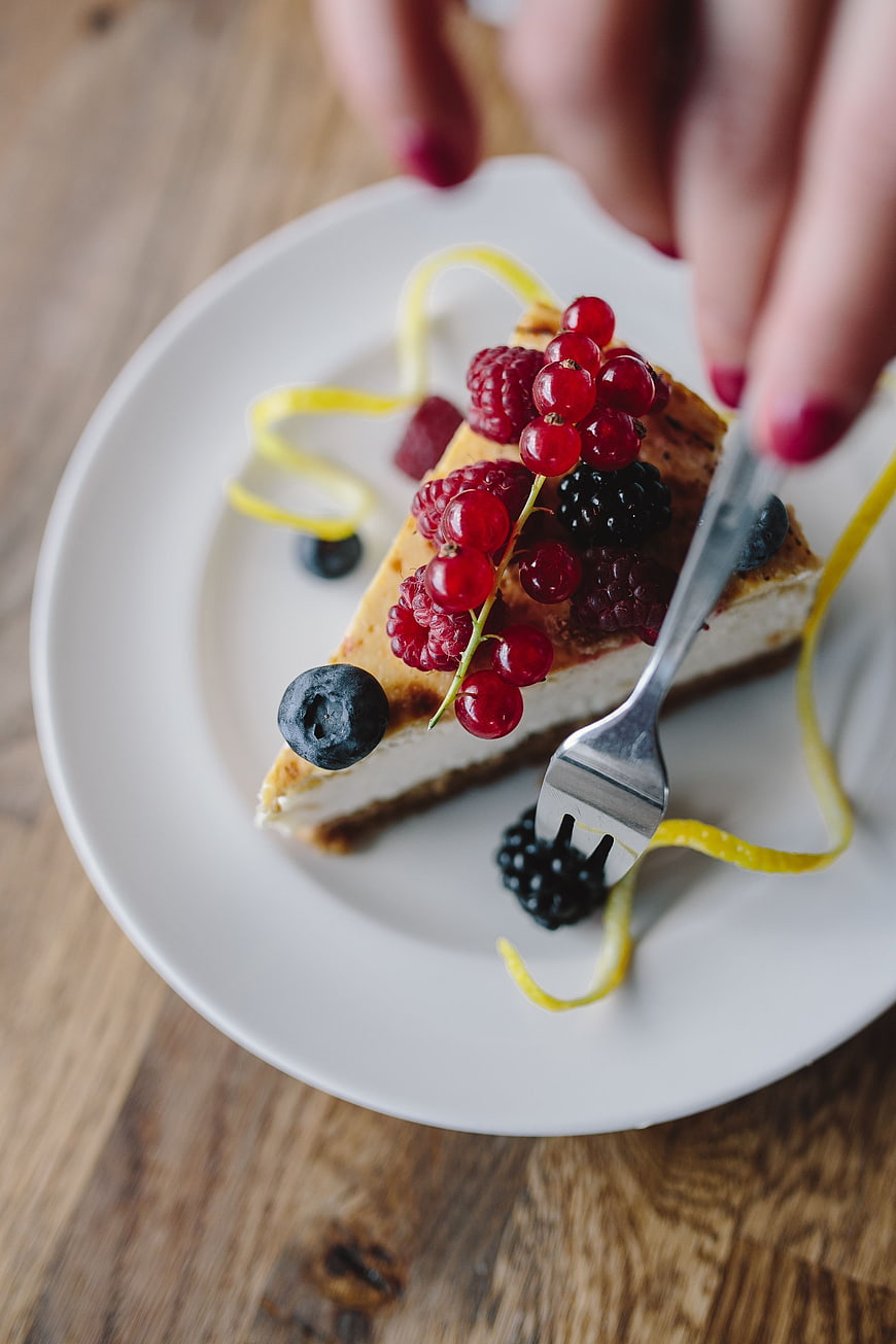 Hand Eating Berry Cheesecake Slice on White Plate