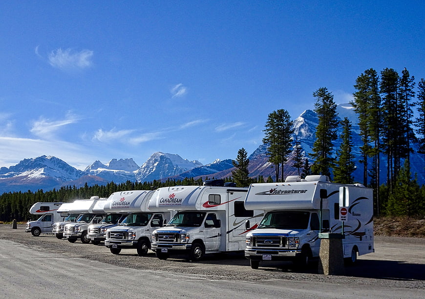 Row of white RVs parked in gravel lot with snow-capped mountains and pine trees