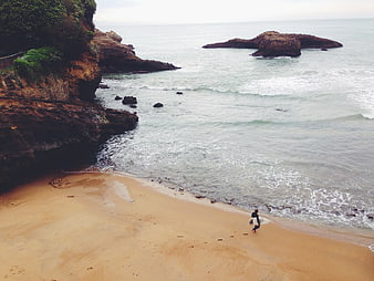 Surfer walking on sandy beach with rocky cliffs and ocean waves