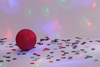 Red glittery ball with confetti on white surface with colorful bokeh lights