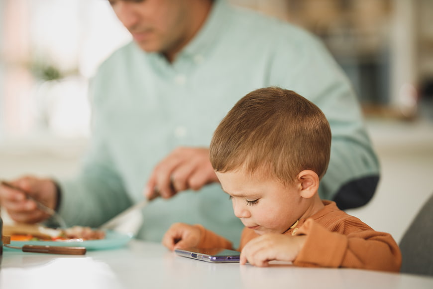 Toddler boy playing with smartphone while father eats breakfast