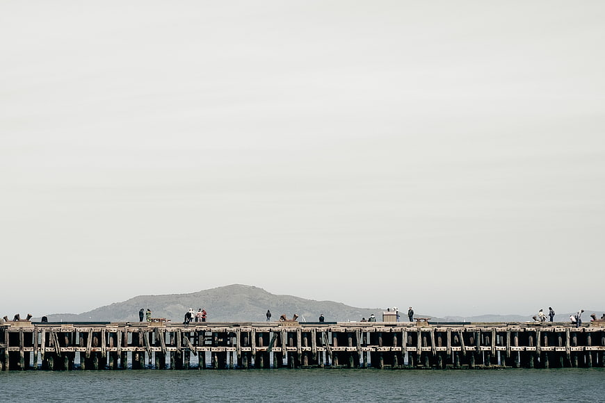 Long wooden pier with people fishing against a mountain backdrop