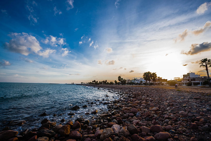 Sunset over Rocky Beach with Coastal Buildings and Blue Sky