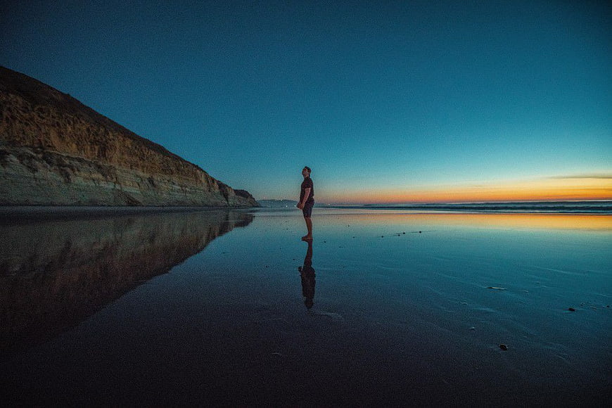 Man Standing on Wet Beach at Sunset with Cliff Reflection