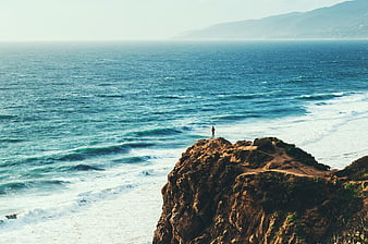 Lone Man Standing on Rocky Cliff Overlooking Ocean Waves