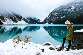 Woman in Green Jacket Sitting by Turquoise Lake in Snowy Mountains
