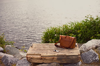 Brown leather bag and smartphone on wooden box by lake