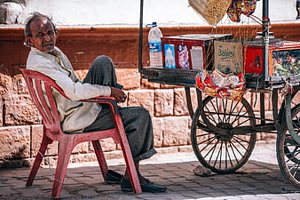 Elderly Indian street vendor sitting on red chair beside cart