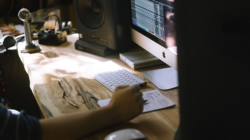 Music Producer Writing Notes at Wooden Desk with Computer