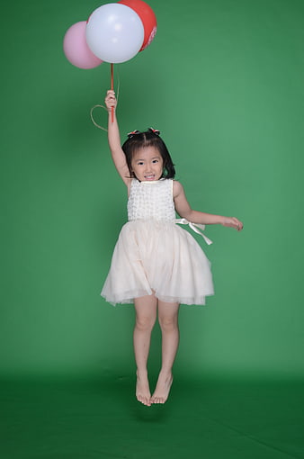Young girl jumping with balloons in white dress against green background