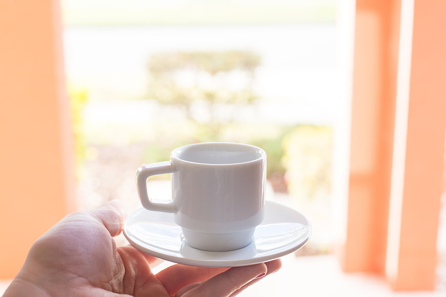 Hand holding white coffee cup on saucer near window