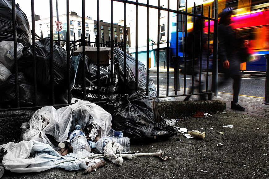 Urban street litter and overflowing trash bags behind metal fence