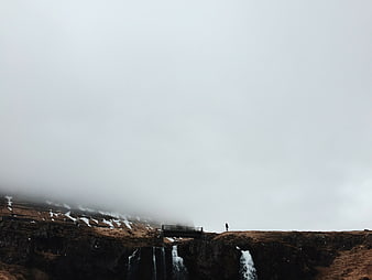 Lone Hiker on Cliff Edge with Waterfalls in Foggy Landscape