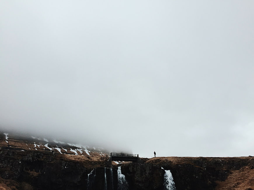 Lone Hiker on Cliff Edge with Waterfalls in Foggy Landscape