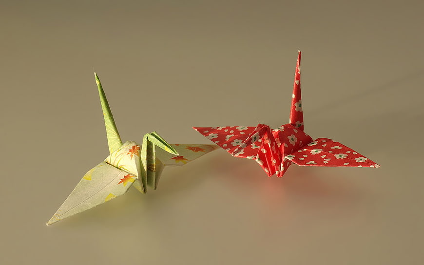Two colorful origami cranes in flight against a neutral beige background
