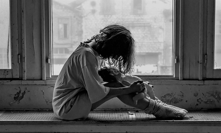 Sad woman sitting by window holding doll in black and white
