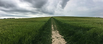 Panoramic view of a dirt path through green fields under cloudy sky