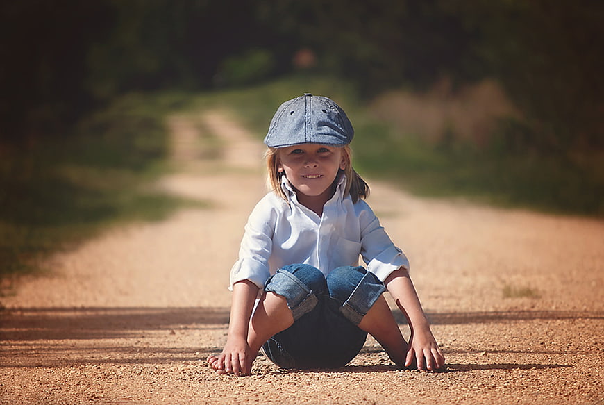Smiling child sitting on a dirt road wearing a cap