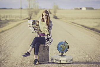 Young woman reading map on suitcase with globe on rural road