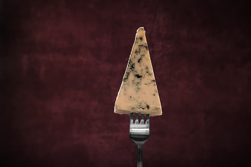 Wedge of blue cheese on a fork against dark red background