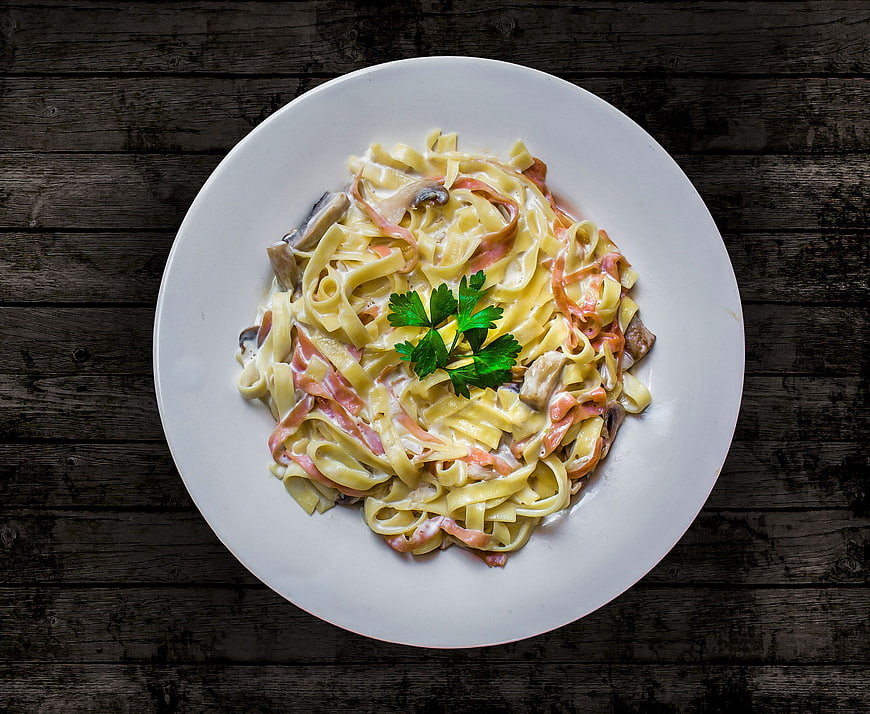 Top View of Tagliatelle Pasta with Ham, Mushrooms, and Parsley on White Plate