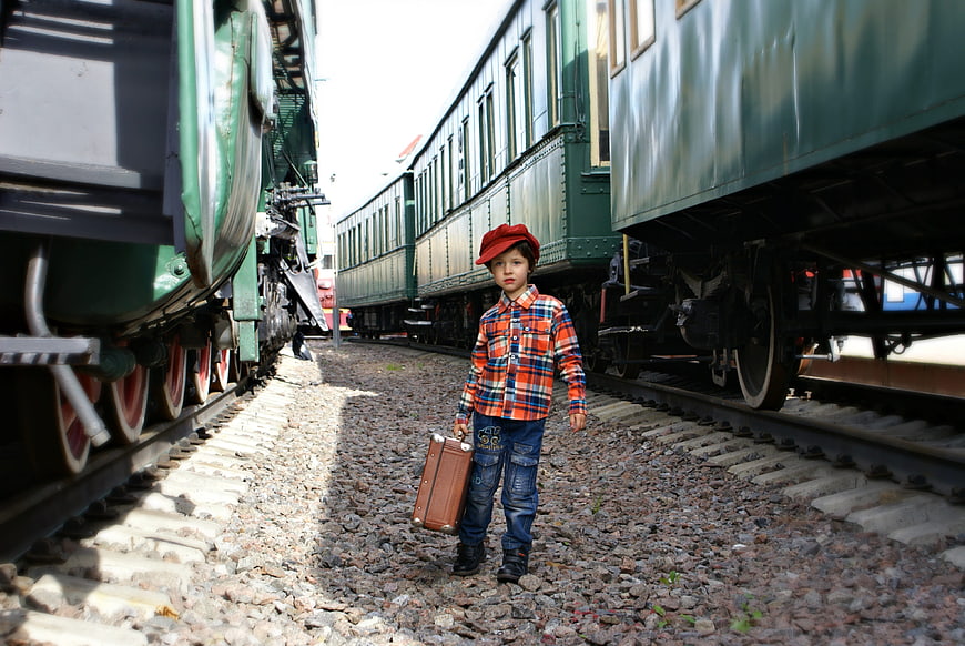 Young boy with vintage suitcase standing between old green trains