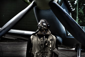 Man in vintage pilot gear and gas mask standing before aircraft propeller