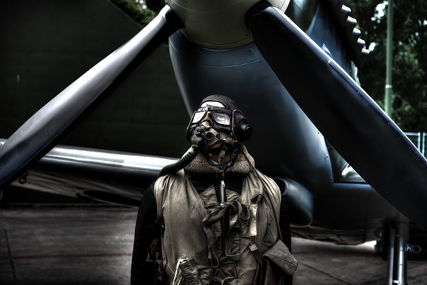 Man in vintage pilot gear and gas mask standing before aircraft propeller