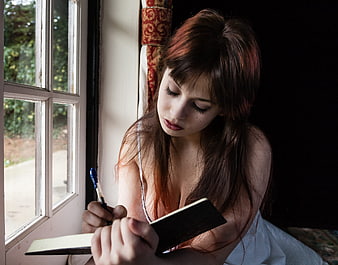 Young woman with red hair writing in a diary by a window