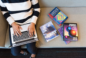 Student typing on laptop with programming textbooks on sofa