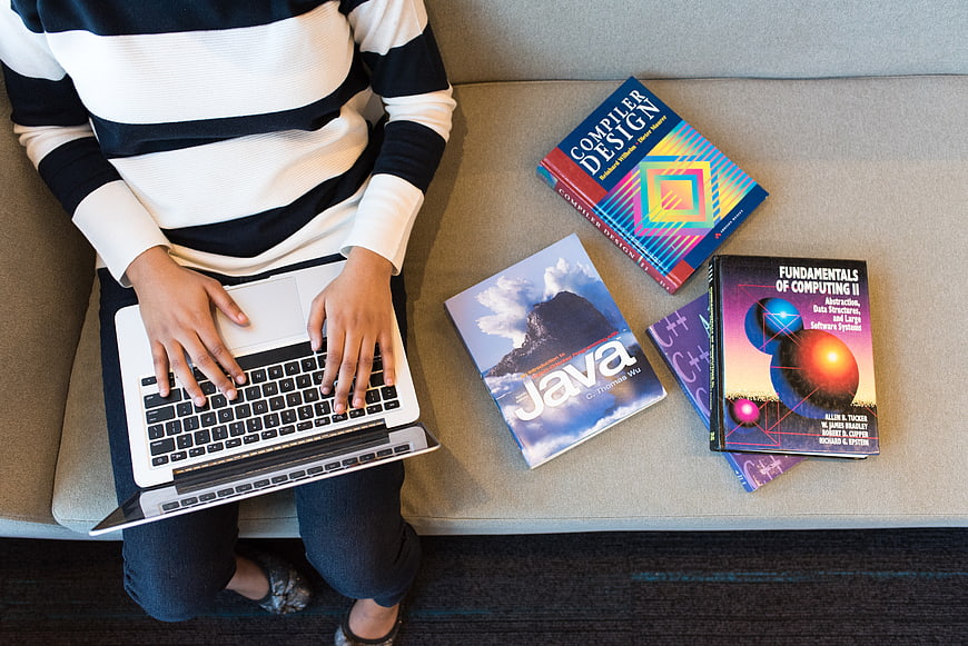 Student typing on laptop with programming textbooks on sofa