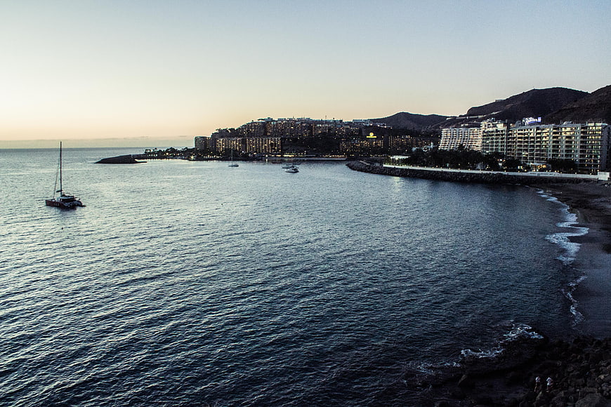 Coastal cityscape at dusk with sailboat and illuminated hotels