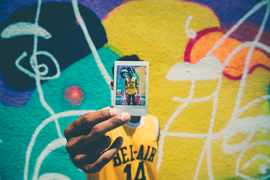 Man holding polaroid photo in front of colorful graffiti wall