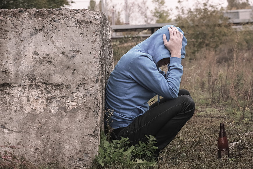 Man in blue hoodie sitting dejectedly against concrete wall outdoors