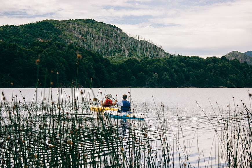 Two people kayaking on a calm lake with forested mountains