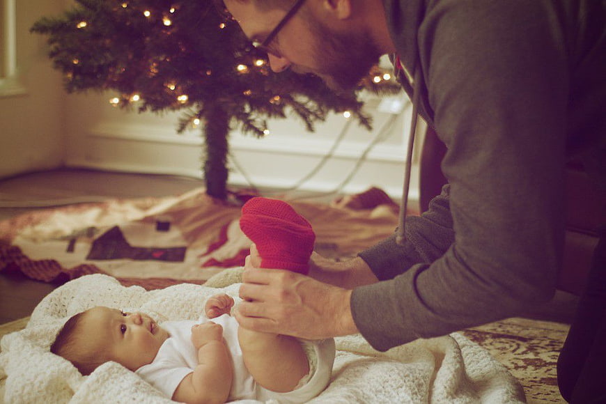 Father Putting Red Sock on Baby Near Christmas Tree
